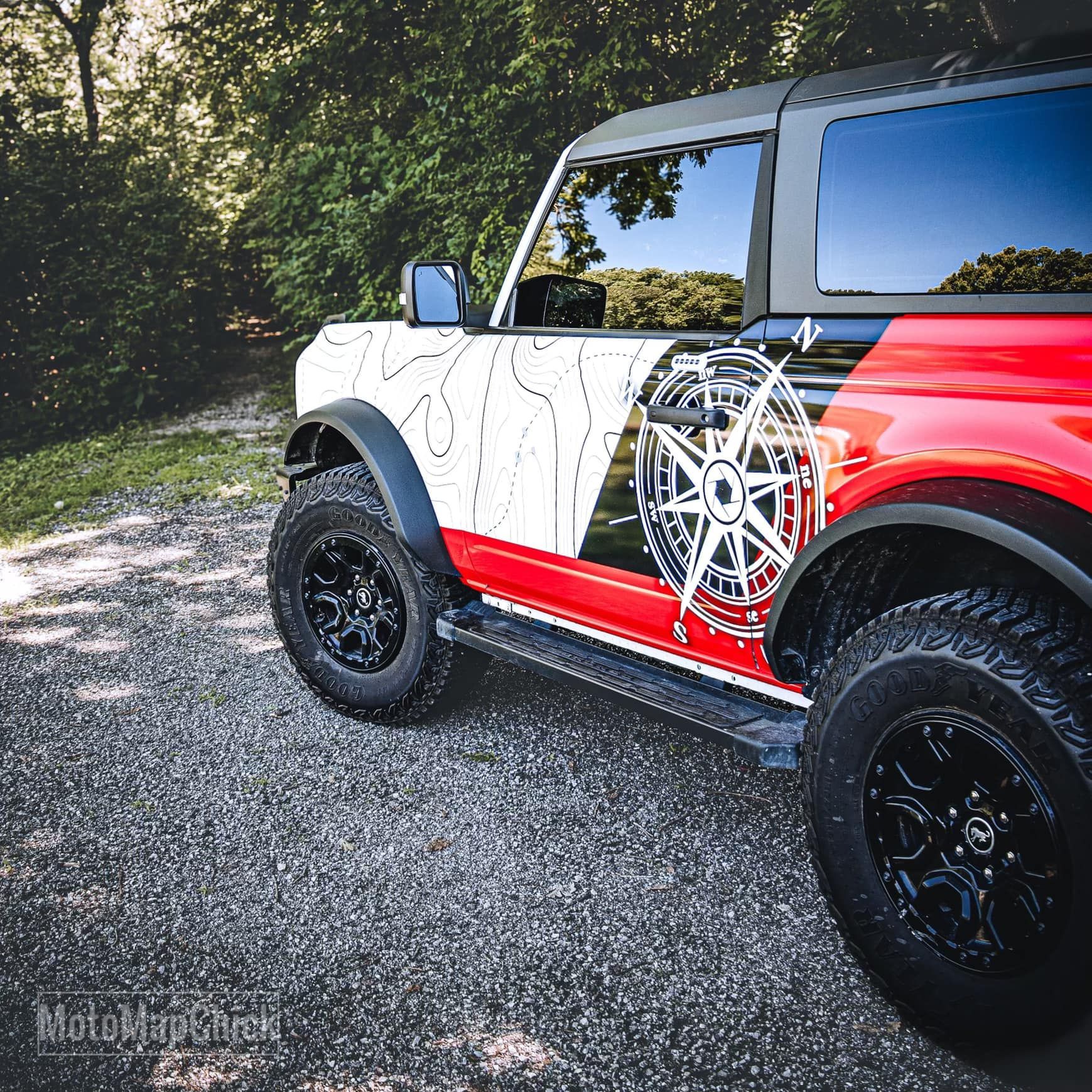 A red and white Ford Bronco with a compass and topographical map decal parked on a gravel path by trees.