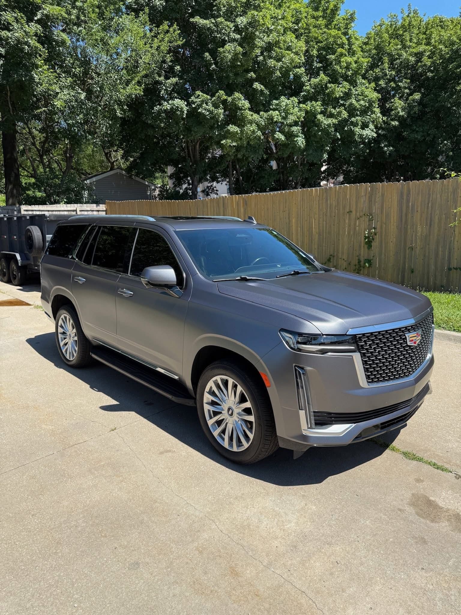 A dark gray Cadillac Escalade SUV parked on a paved driveway in front of a wooden fence and green trees.