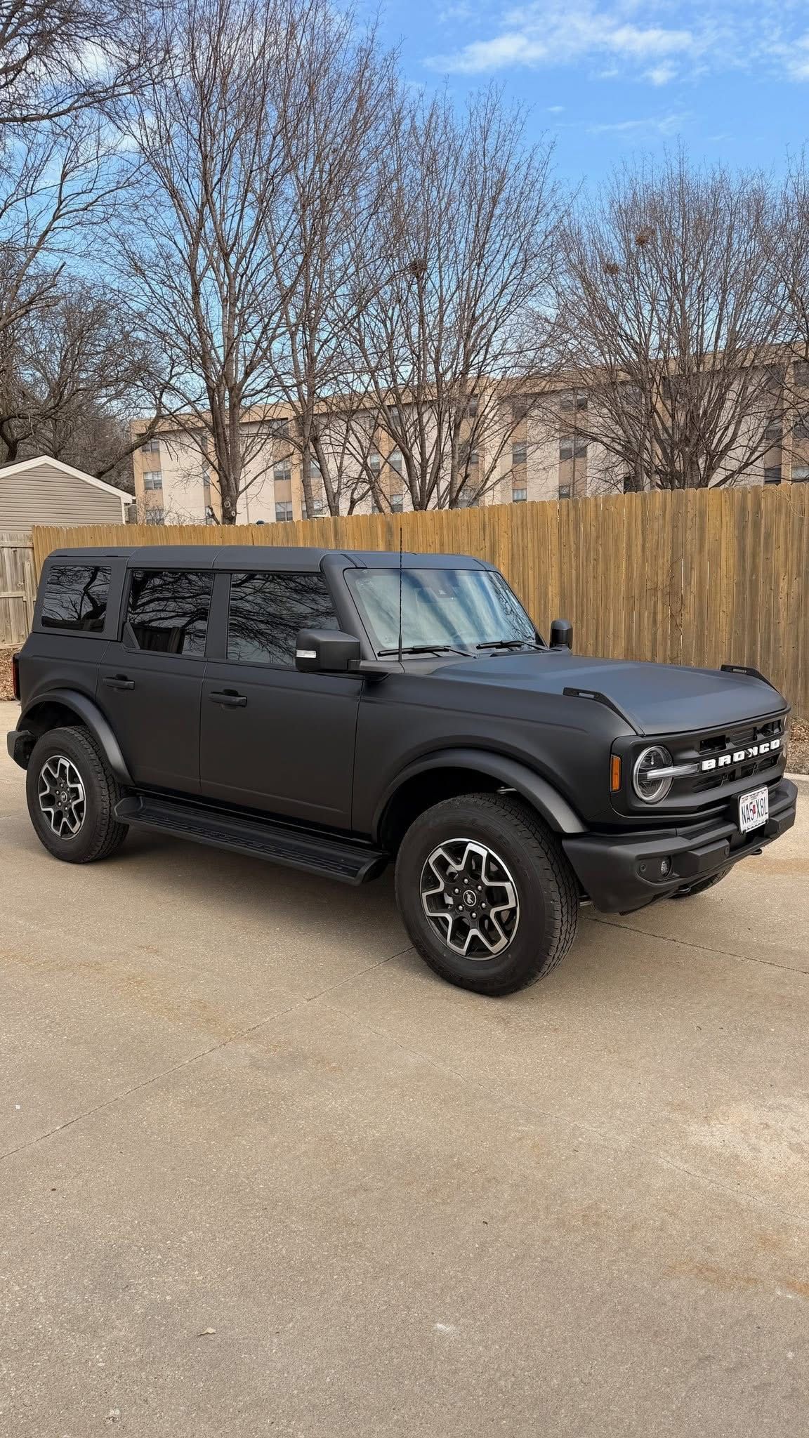 A matte black Ford Bronco SUV parked on a gravel surface in front of a wooden fence and bare trees.