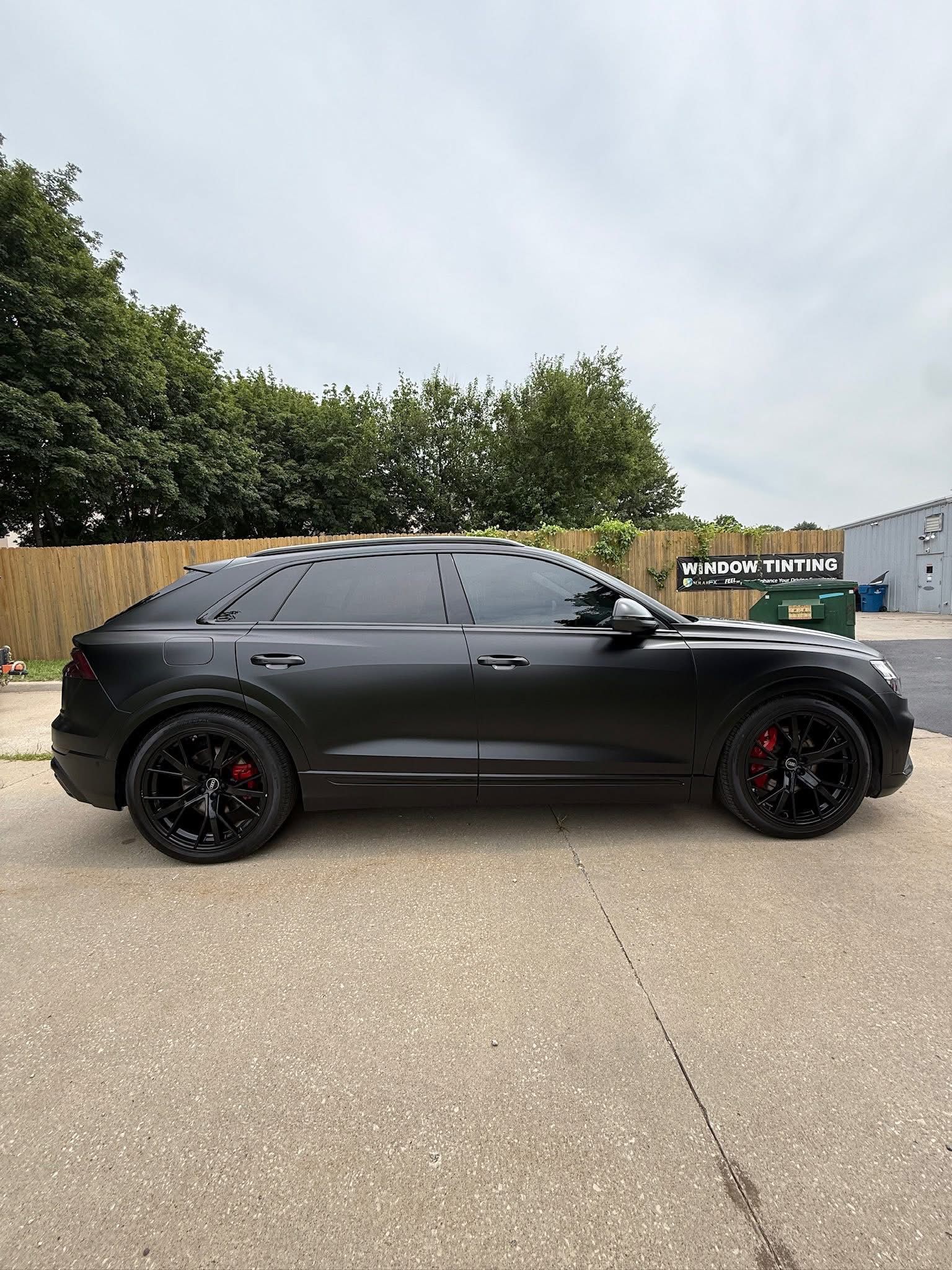 A matte black Audi RS Q8 SUV parked on an asphalt lot in front of trees and a building under a cloudy sky.