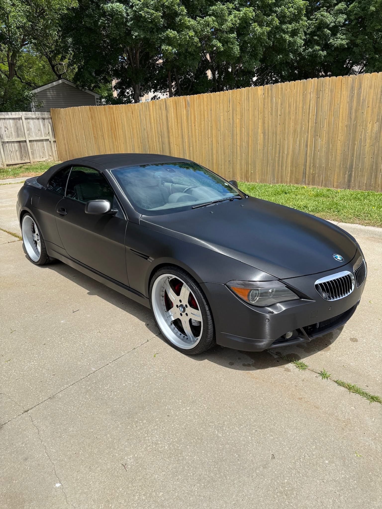 A matte black BMW 6 series convertible with chrome rims parked on a concrete driveway in front of a wooden fence.