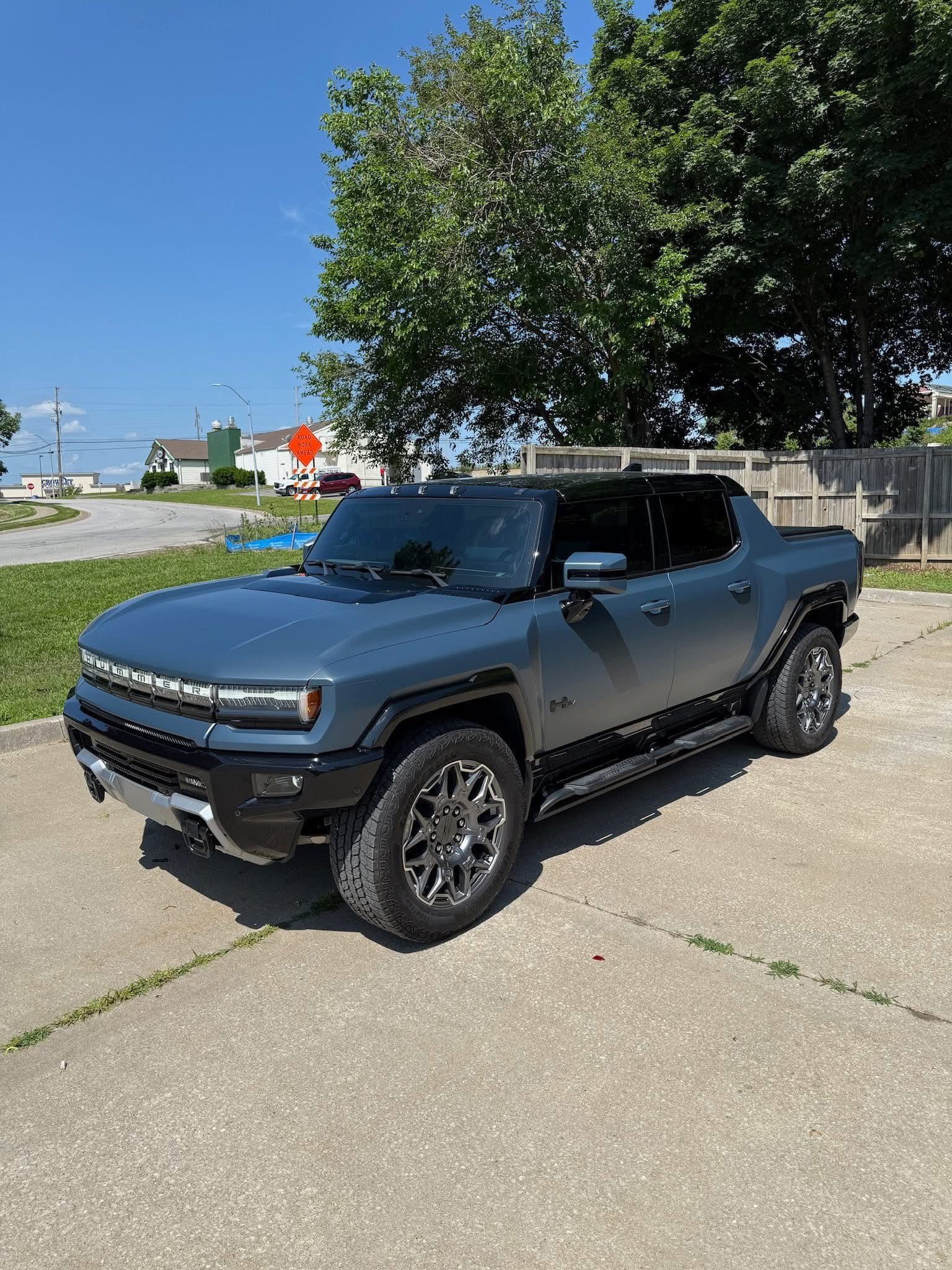 A dark gray Hummer EV pickup truck parked on a concrete lot under a clear blue sky with trees in the background.