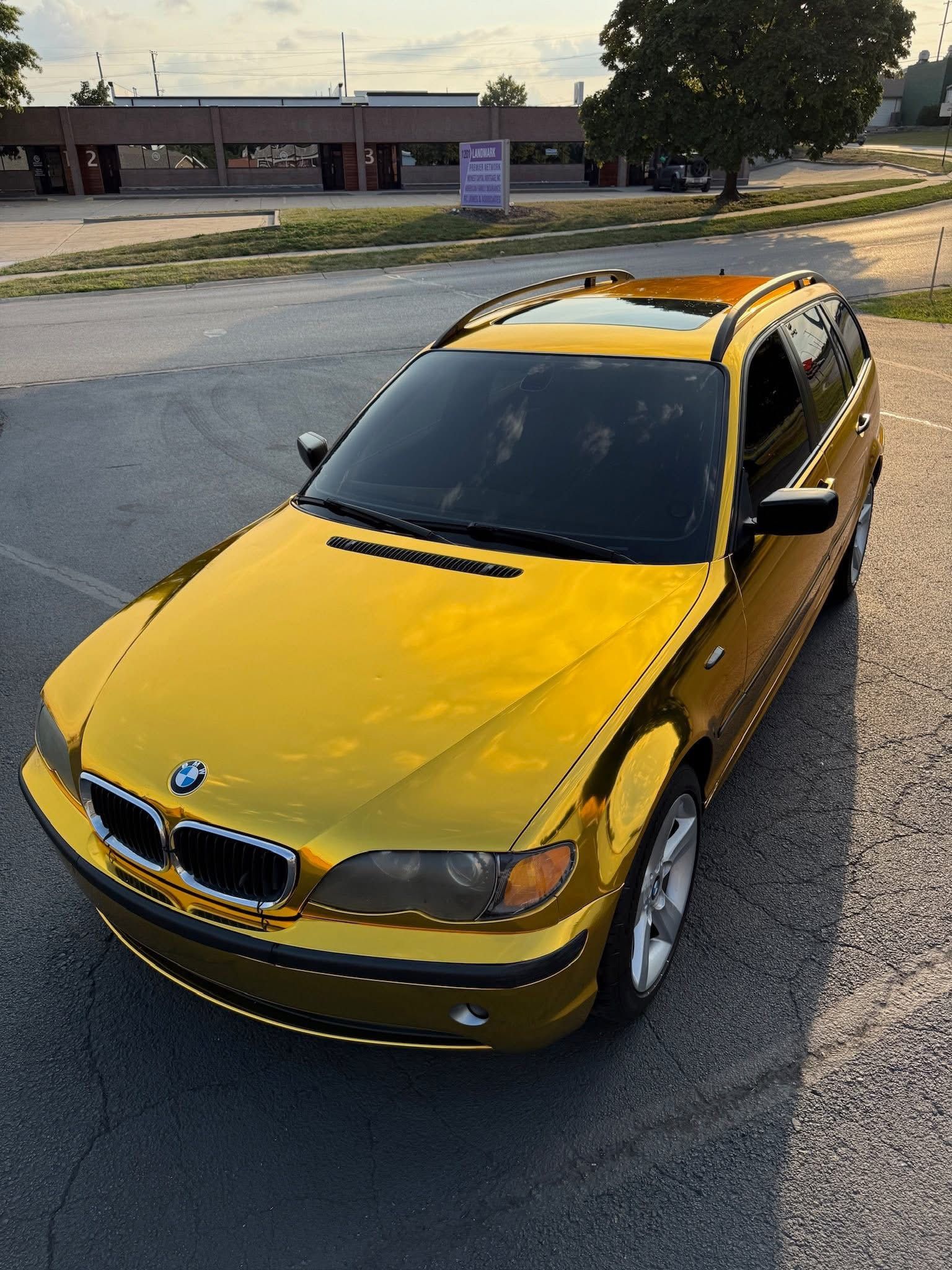 A gold chrome-wrapped BMW station wagon parked on an asphalt lot during sunset.