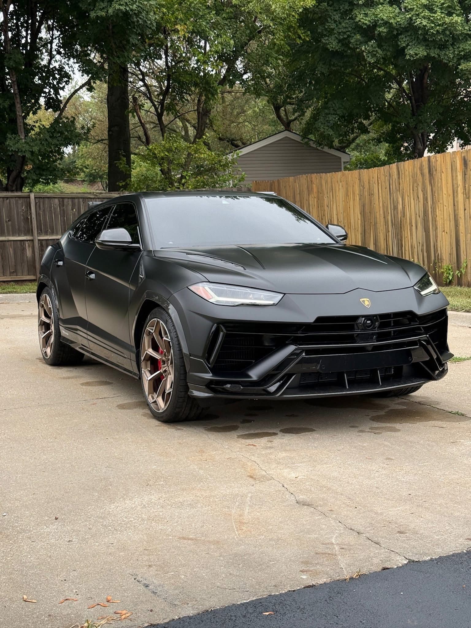 A matte black Lamborghini Urus SUV parked on a concrete driveway in front of a wooden fence and trees.