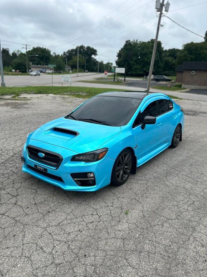 A bright turquoise Subaru WRX with black wheels and a black roof, parked in an outdoor lot on a cloudy day.
