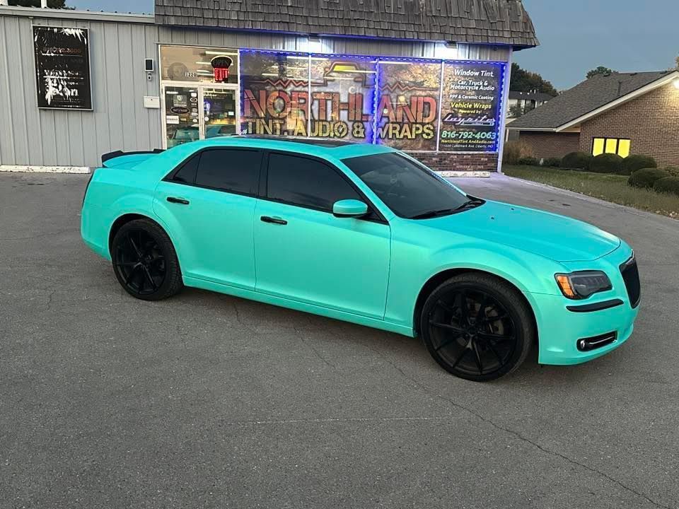 A bright turquoise sedan with black rims parked in front of a storefront advertising vehicle wraps.