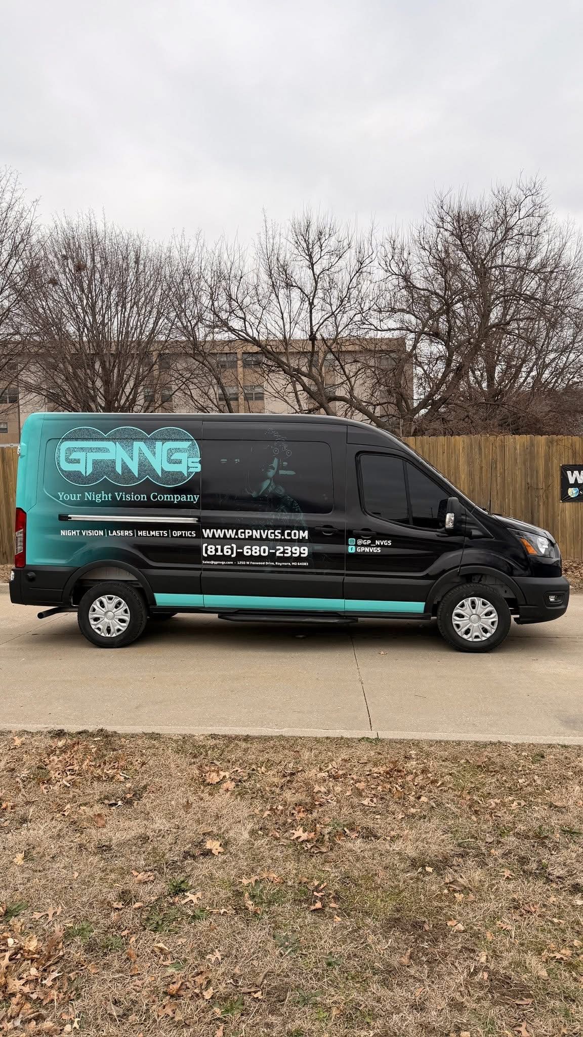 A black and teal commercial cargo van parked outdoors on a paved lot in front of a wooden fence and leafless trees.