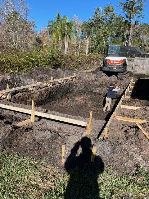 Construction site: Excavator and worker building a structure, with wooden frames, in a dirt backyard on a sunny day.