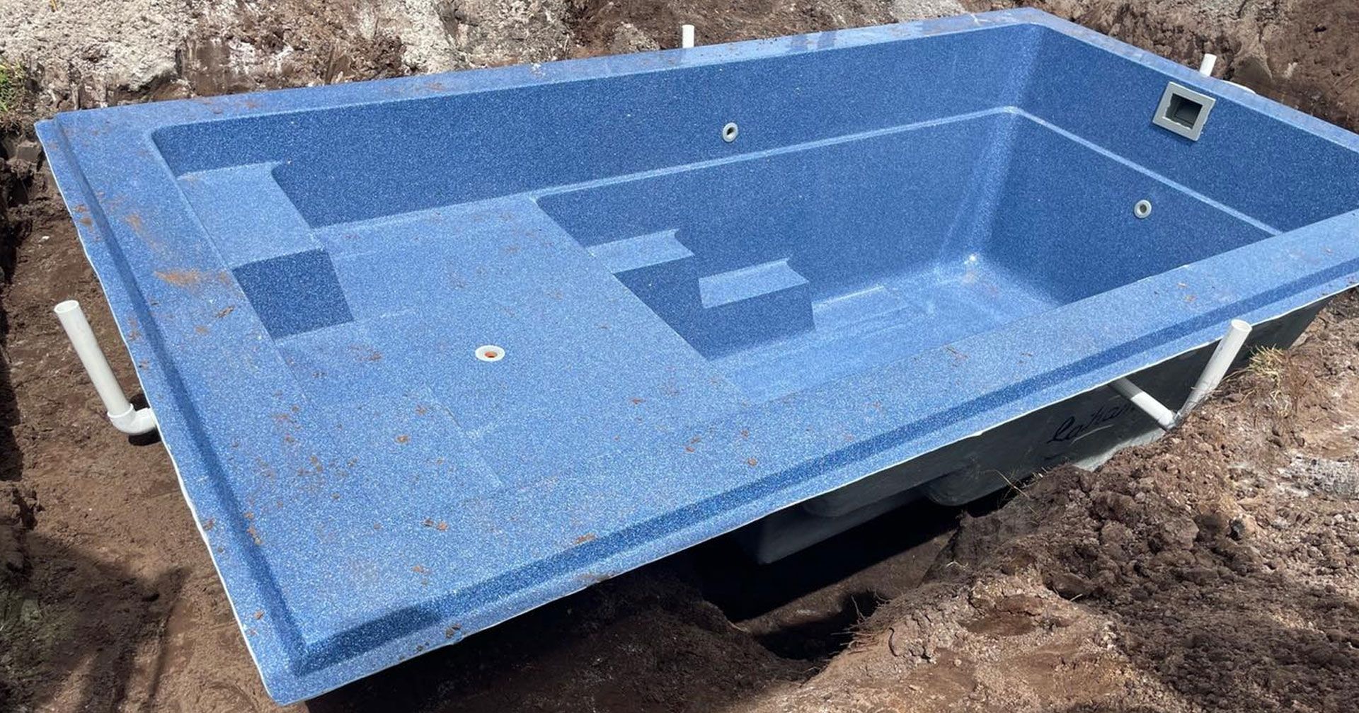 Blue rectangular pool shell in an excavated hole, with plumbing visible.