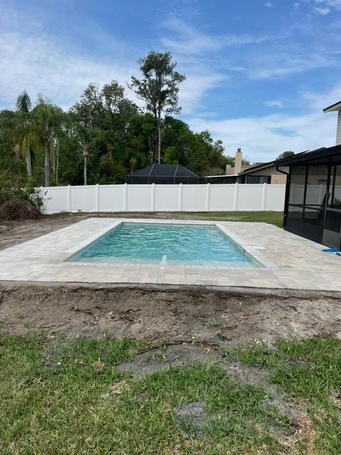 Rectangular swimming pool with light-colored pavers, surrounded by a white fence and green lawn under a blue sky.