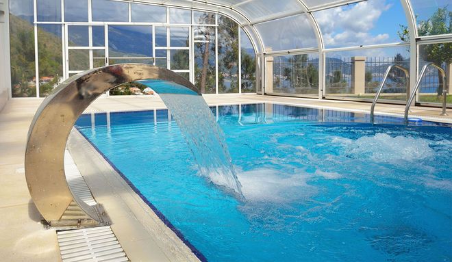 Swimming pool with a water feature, under a glass enclosure, with a scenic outdoor view.