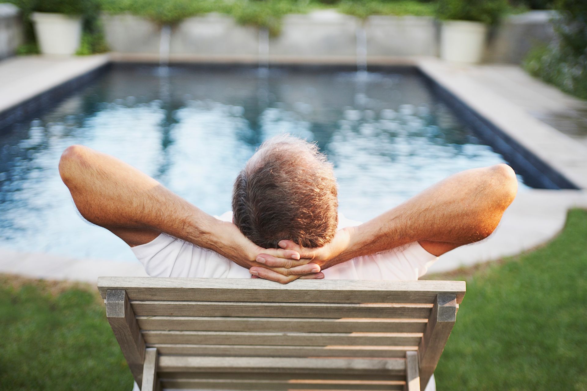 Man relaxing in a chair by a pool, arms behind head.