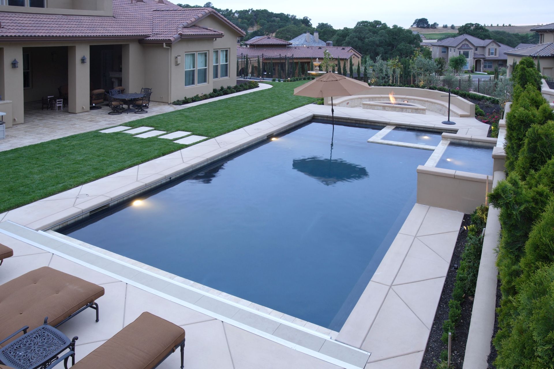 Backyard pool with tan patio, lawn, house, and landscaping. Dark blue water.