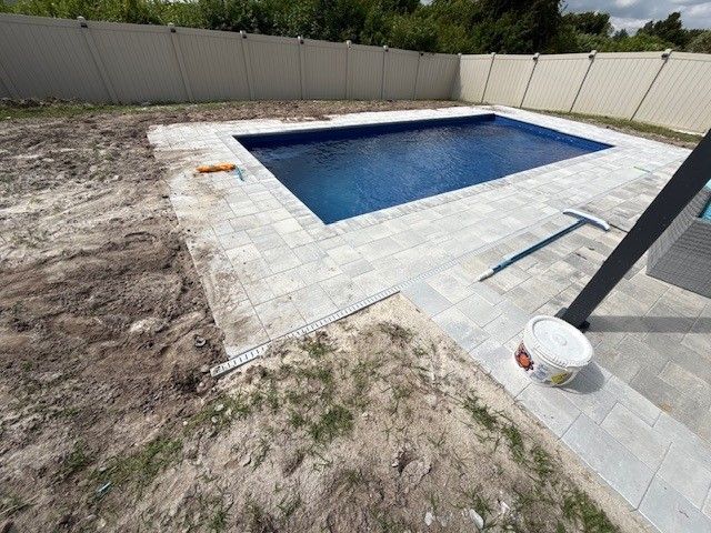 A rectangular swimming pool surrounded by gray pavers, fence, and dirt.