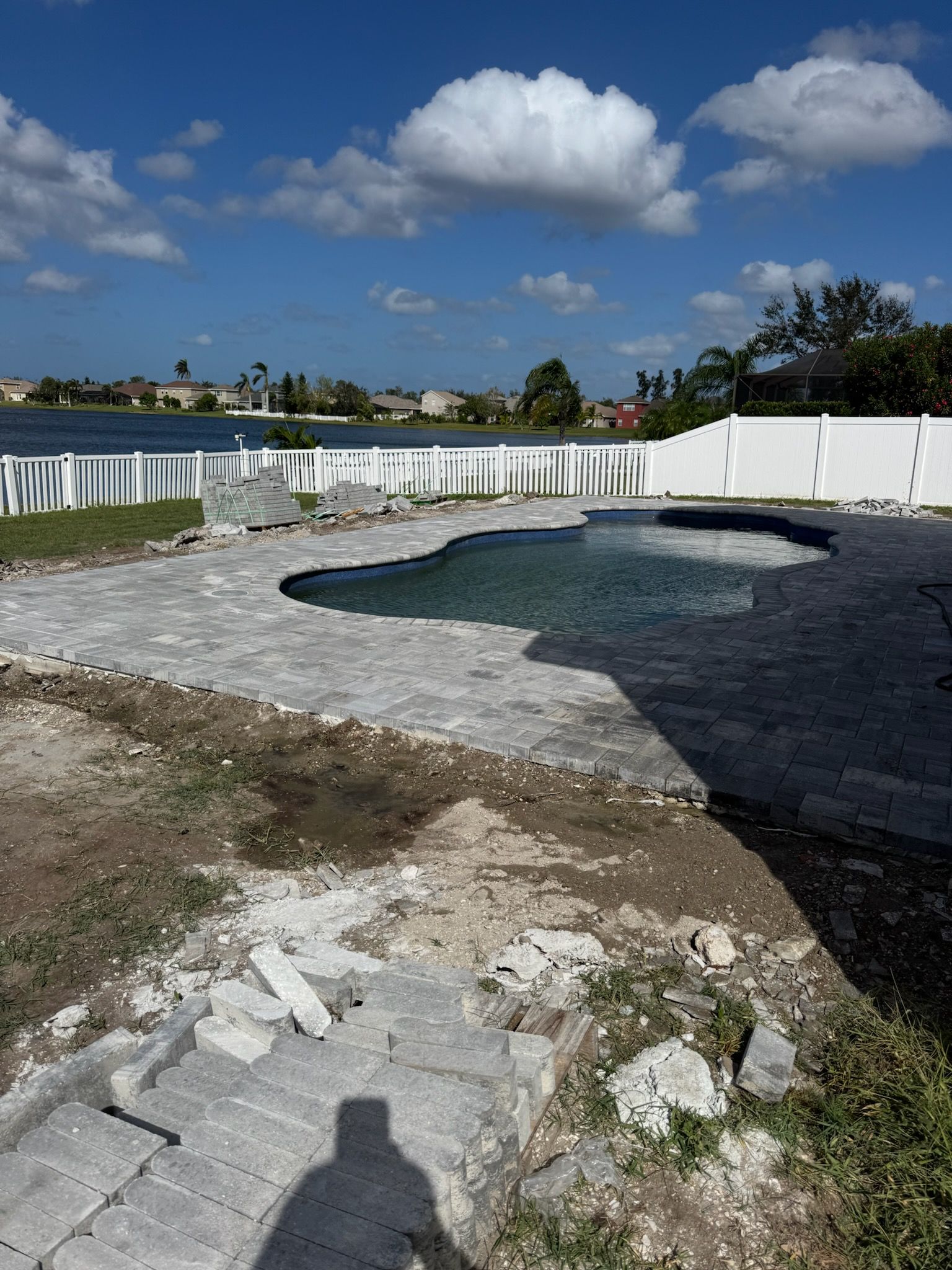 Backyard with a pool, paved patio, and white fence overlooking a lake on a sunny day.