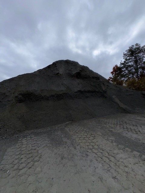 A large pile of dark gray gravel under a cloudy sky. Tire tracks in the foreground.