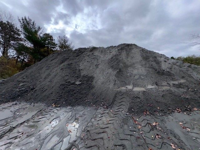 Large pile of gray gravel with tire tracks on muddy ground, under a cloudy sky.