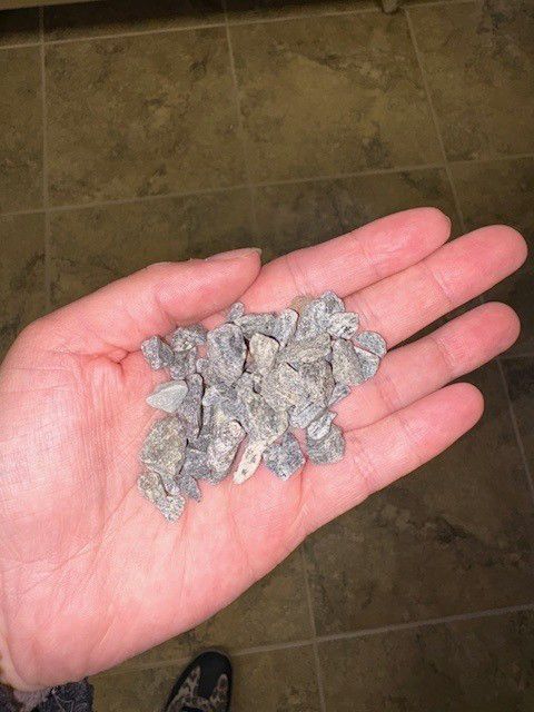 Hand holding a pile of small, grey rocks. Brown tile floor in background.