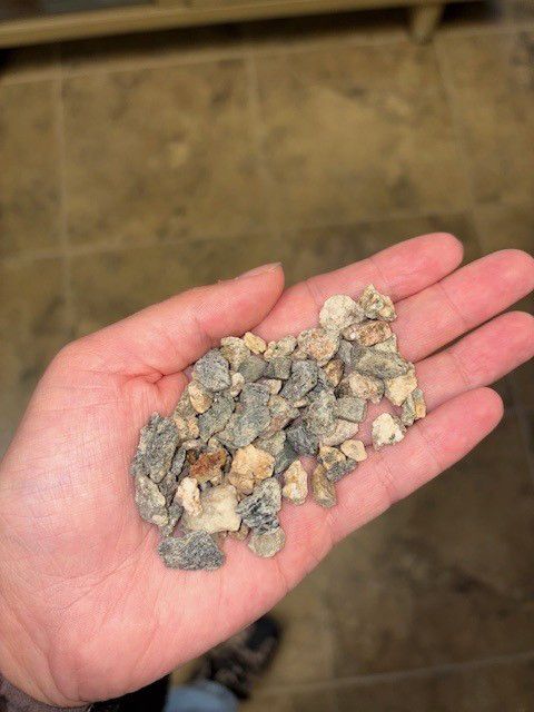 Hand holding small, irregular pieces of gray and beige gravel, over a tiled floor.