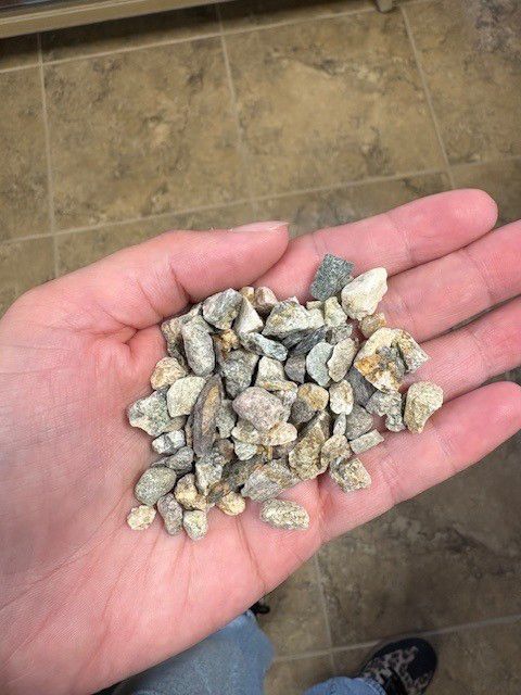 Handful of small, light-colored gravel stones against a skin tone hand, brown tiled floor visible.