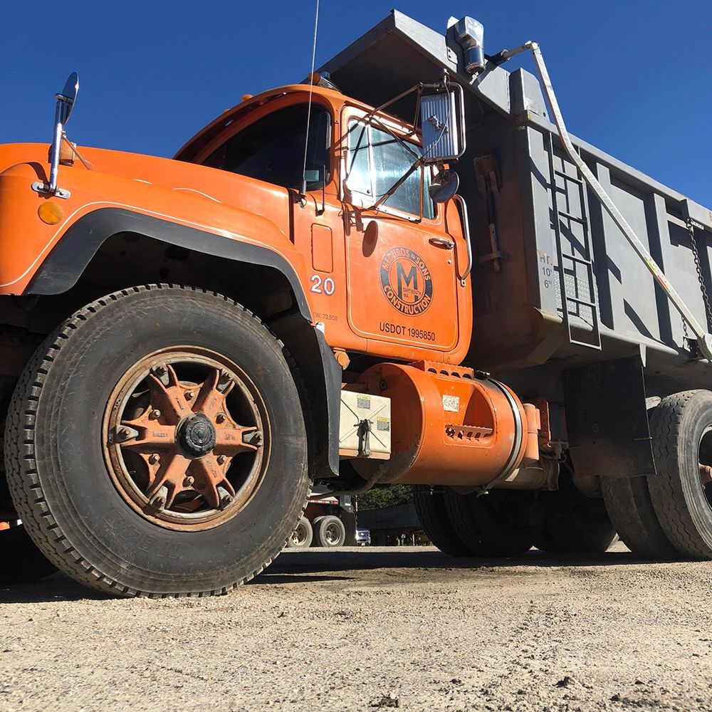 Orange Mack dump truck with a raised bed, parked on a gravel surface. Clear blue sky in the background.