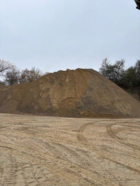 Large pile of tan gravel and dirt against a cloudy sky. Tire tracks in the foreground.