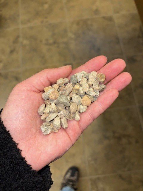 Hand holding a pile of small, light-colored gravel against a neutral floor background.