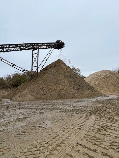 A large pile of sand at a quarry, with a conveyor belt overhead, under a cloudy sky.