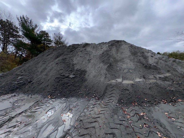 Pile of gray gravel with tire tracks, trees in the background, under a cloudy sky.