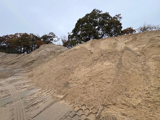 Large pile of beige sand with tire tracks in the foreground, trees on the horizon under a cloudy sky.