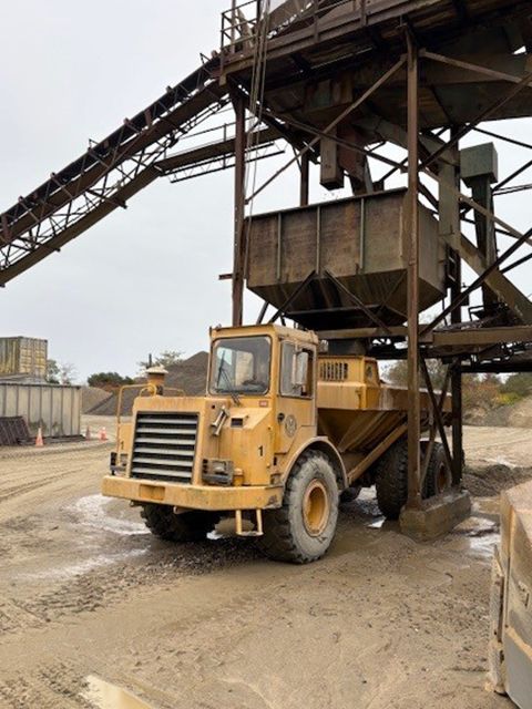 Yellow dump truck under a metal structure, likely at a quarry. Overcast sky.
