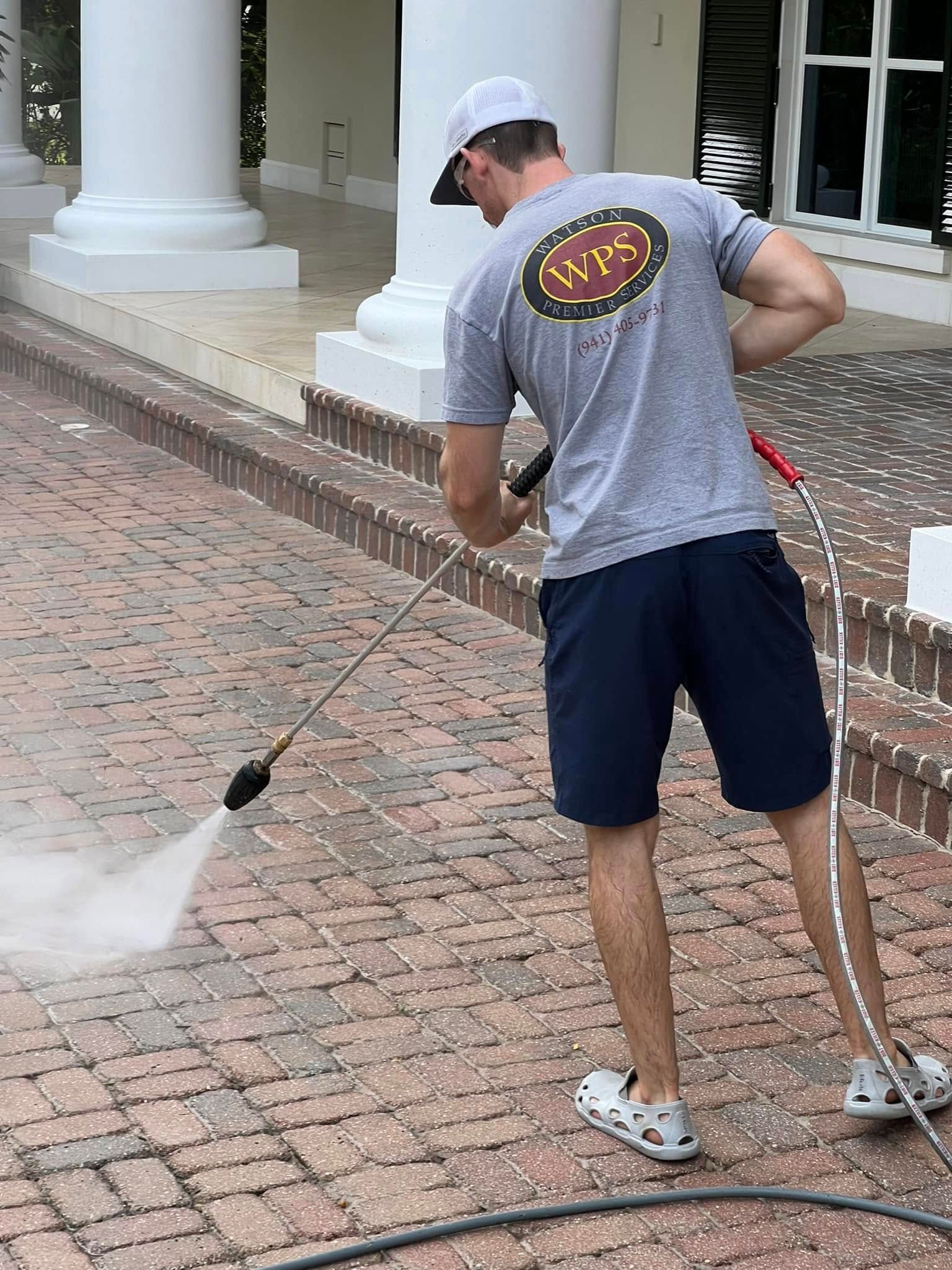 Man power washing brick walkway with high-pressure hose, in front of a white building with columns.