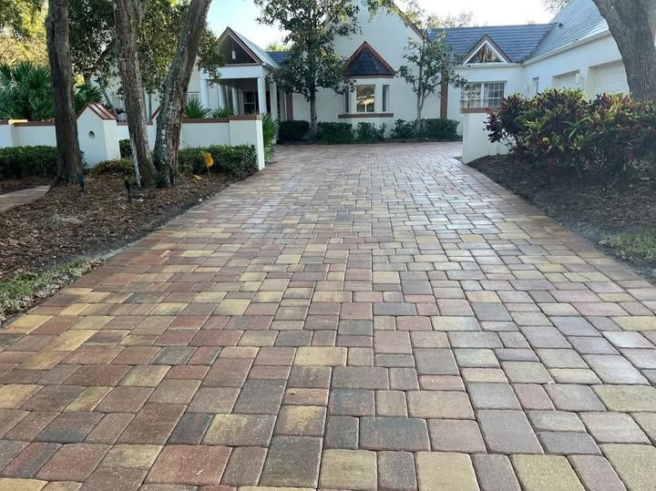 Brick paver driveway leading to a white house with a landscaped yard and trees.