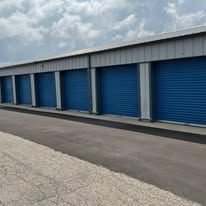 A row of closed blue metal roll-up doors on a self-storage building with a gray asphalt driveway in front.