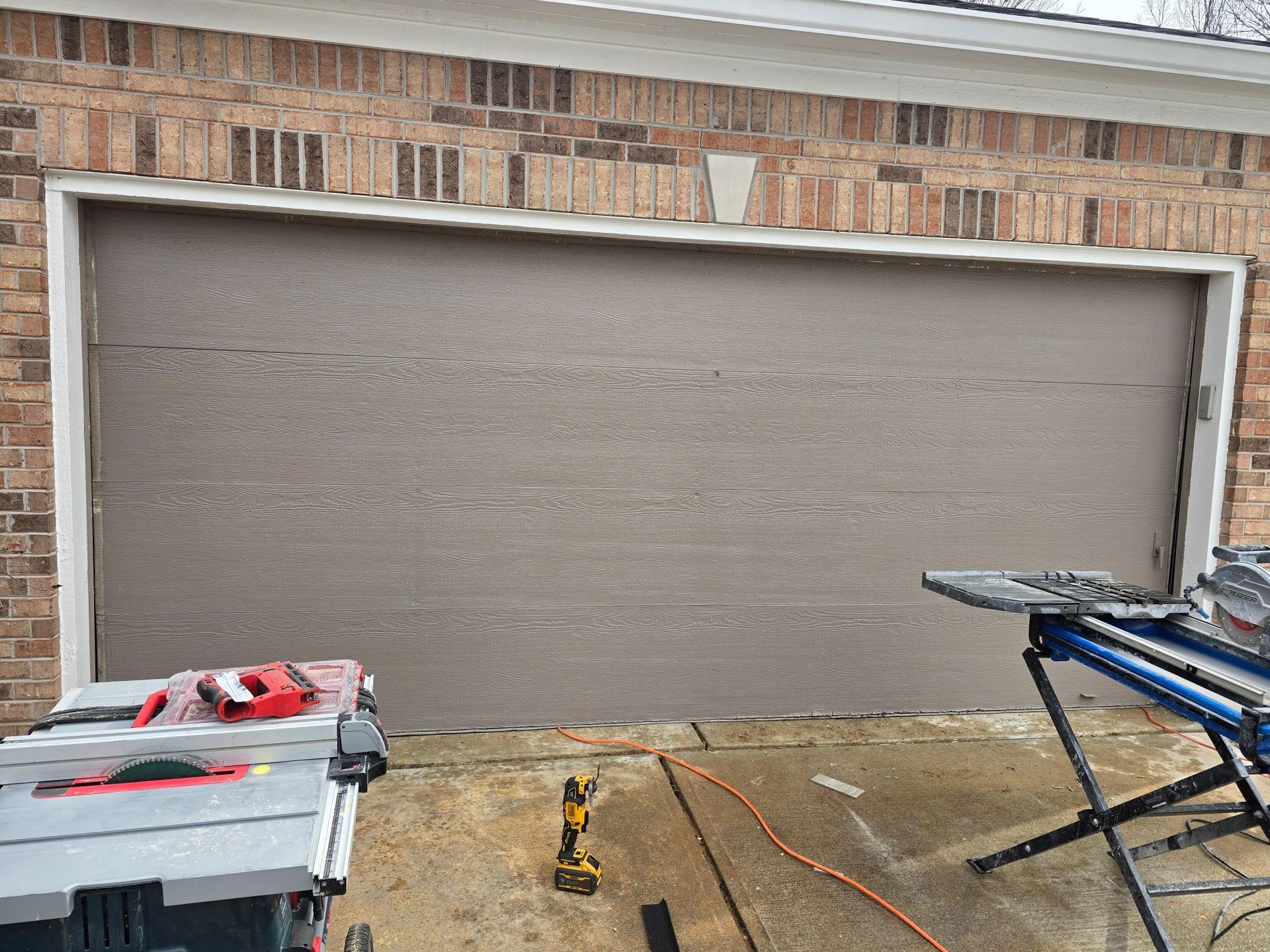 A beige garage door installed in a brick house, with two power tools on stands in the foreground.