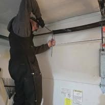 A person in dark work clothes standing on a ladder to repair a garage door spring.