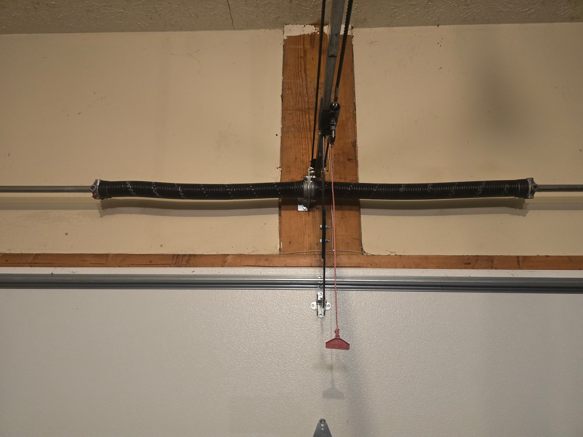 A horizontal garage door torsion spring mounted on a wooden header above a white garage door.