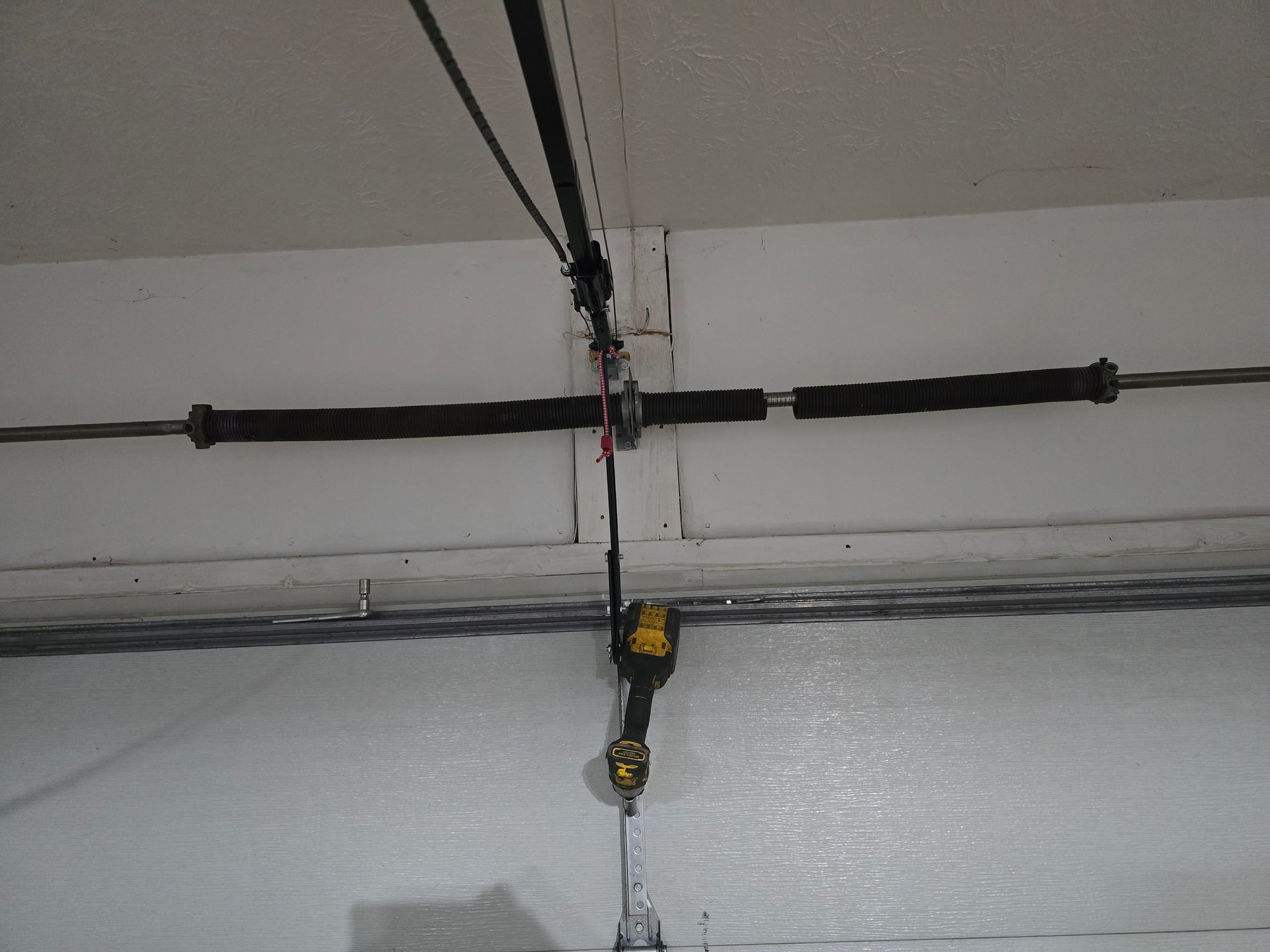 A black and yellow cordless drill hangs by a cord from a garage door torsion spring assembly mounted on a white wall.