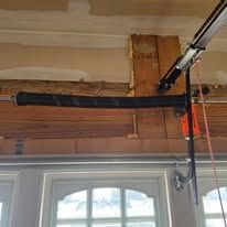 A close-up view of a black garage door torsion spring mounted on a wooden header above a window.