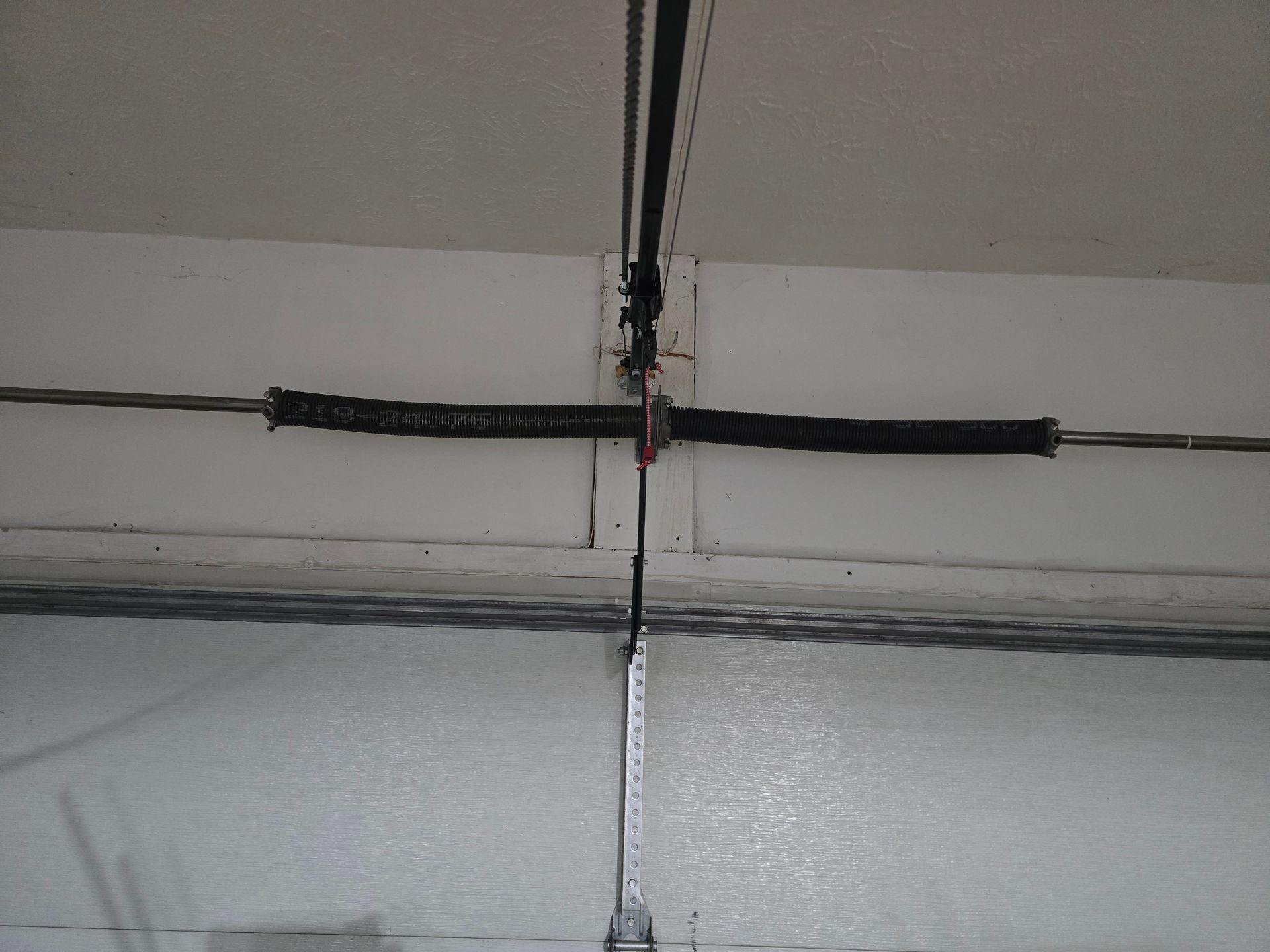 A black, horizontal metal spring mounted above a garage door, centered against a white wall.