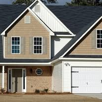 A two-story suburban home with tan siding, a brick lower level, dark roof shingles, and a white two-car garage.