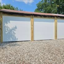 A white, three-bay pole barn structure with wood framing and a metal roof, situated above a gravel driveway.