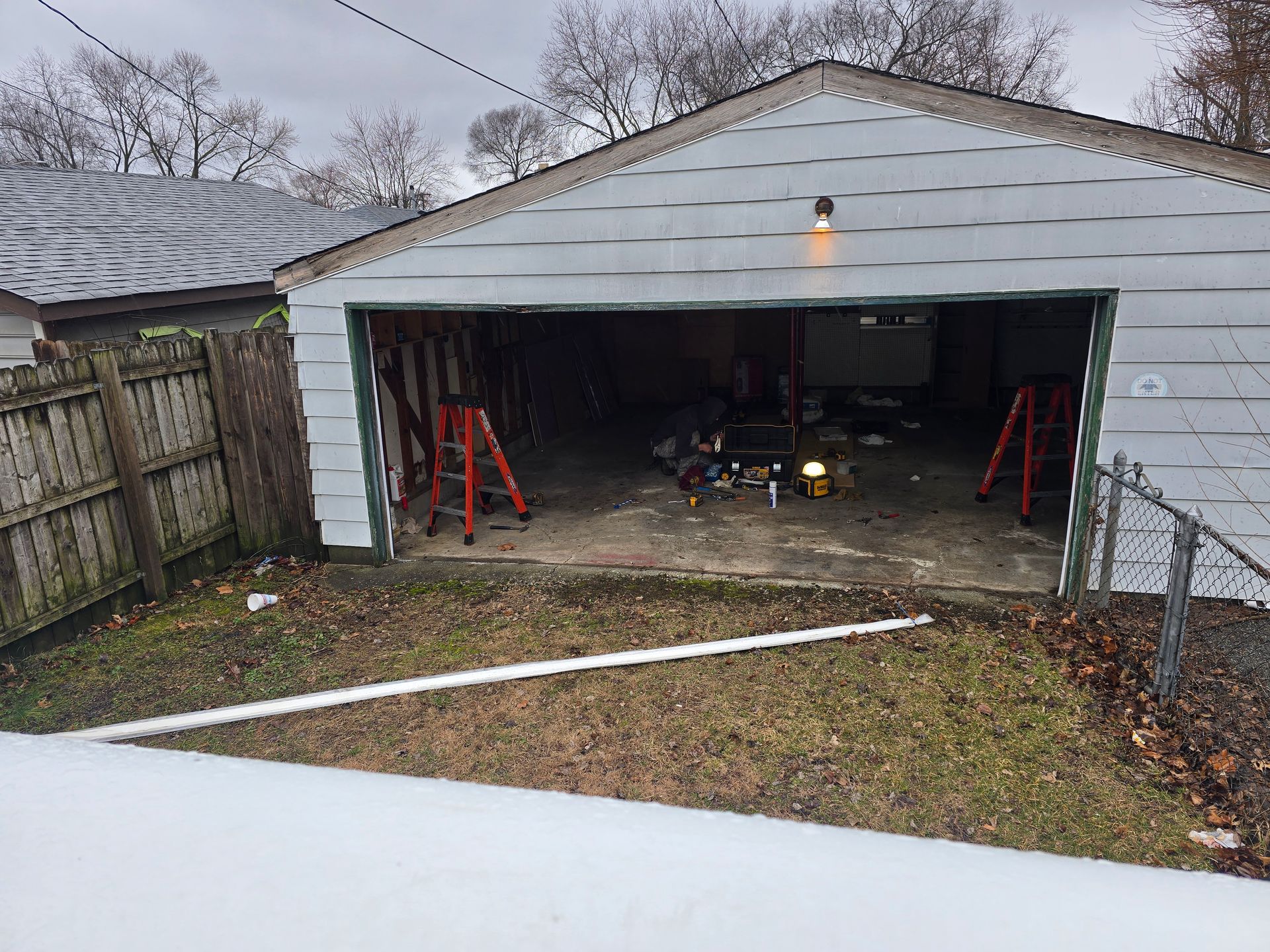 A white garage with an open door stands at the end of a yard, featuring two red ladders inside and a nearby fence.