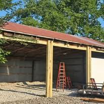 An open-sided wooden structure with a red roof, gravel ground, and a red ladder leaning against the back wall.