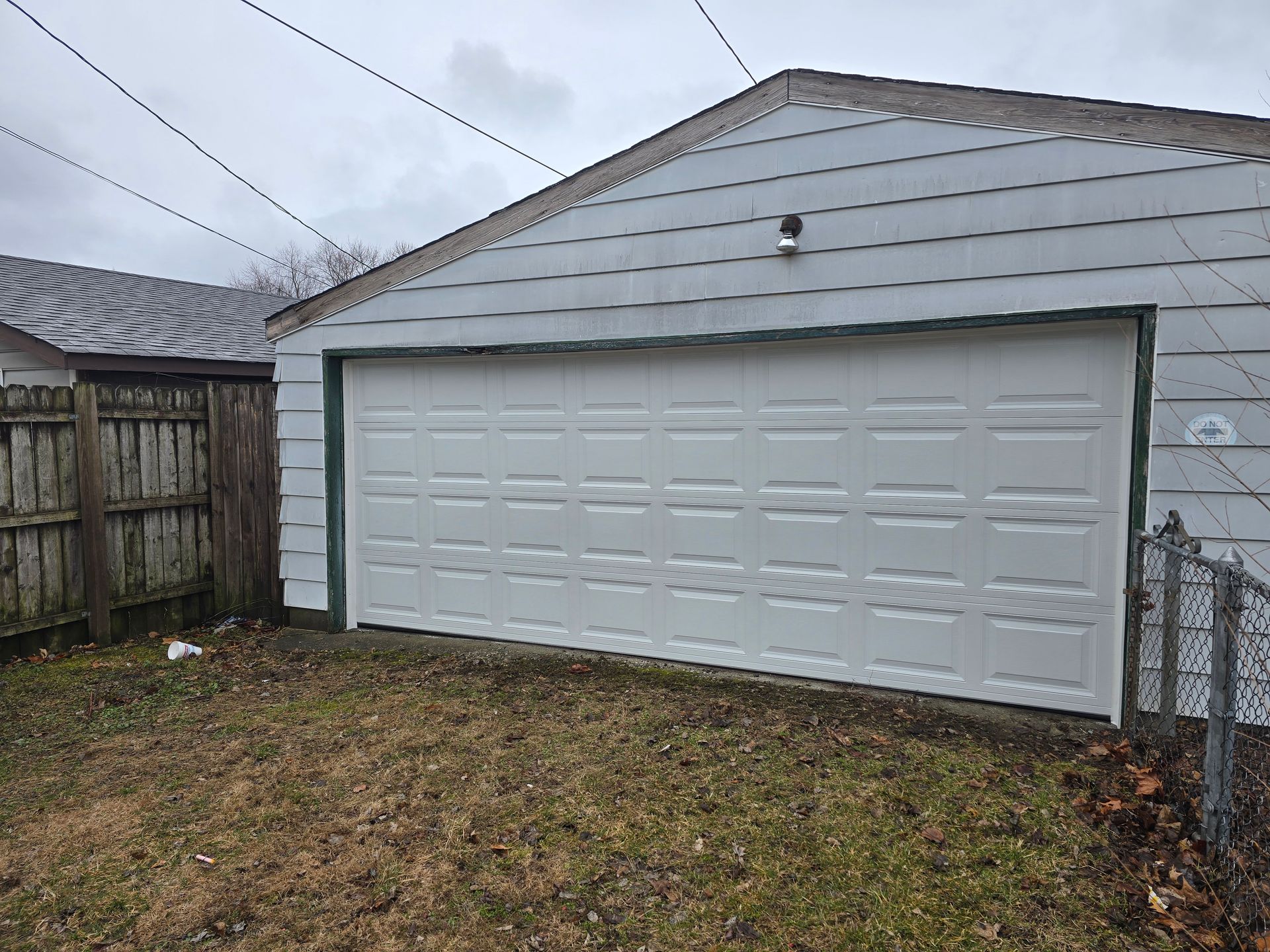 A white garage door on a gray-sided building next to a wooden fence and a chain-link fence on a grassy lot.