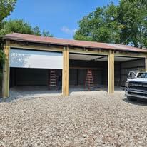 A three-bay wooden garage structure with a gravel driveway, one white door installed, and a white truck parked to the side.