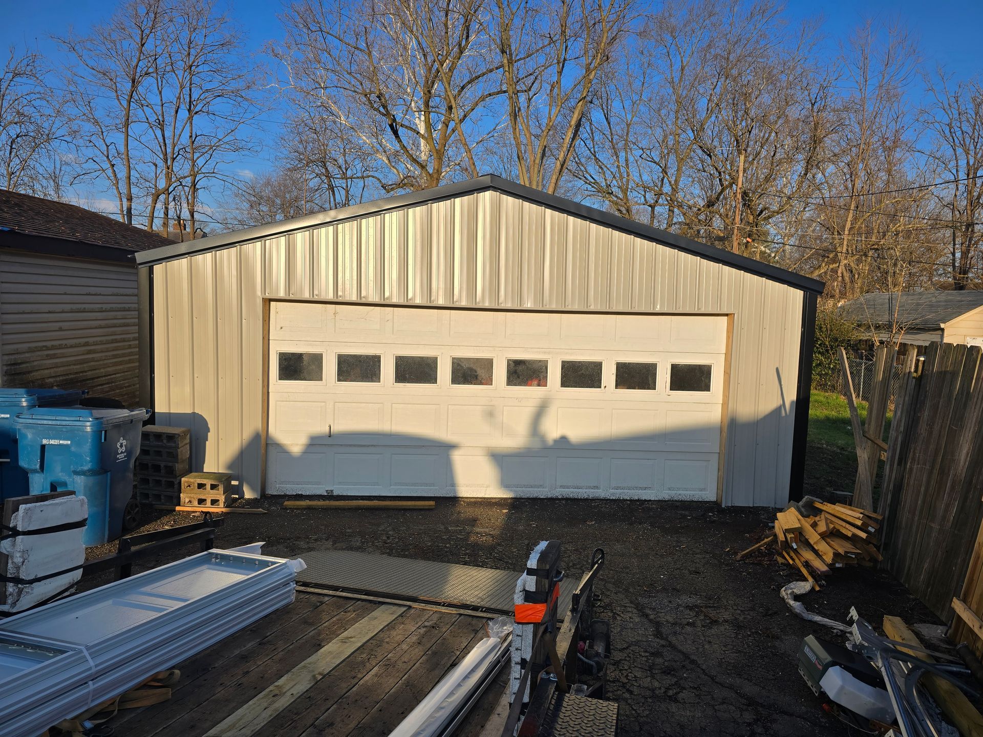 A light-colored metal detached garage with a white, multi-pane windowed door, surrounded by an outdoor area with debris.
