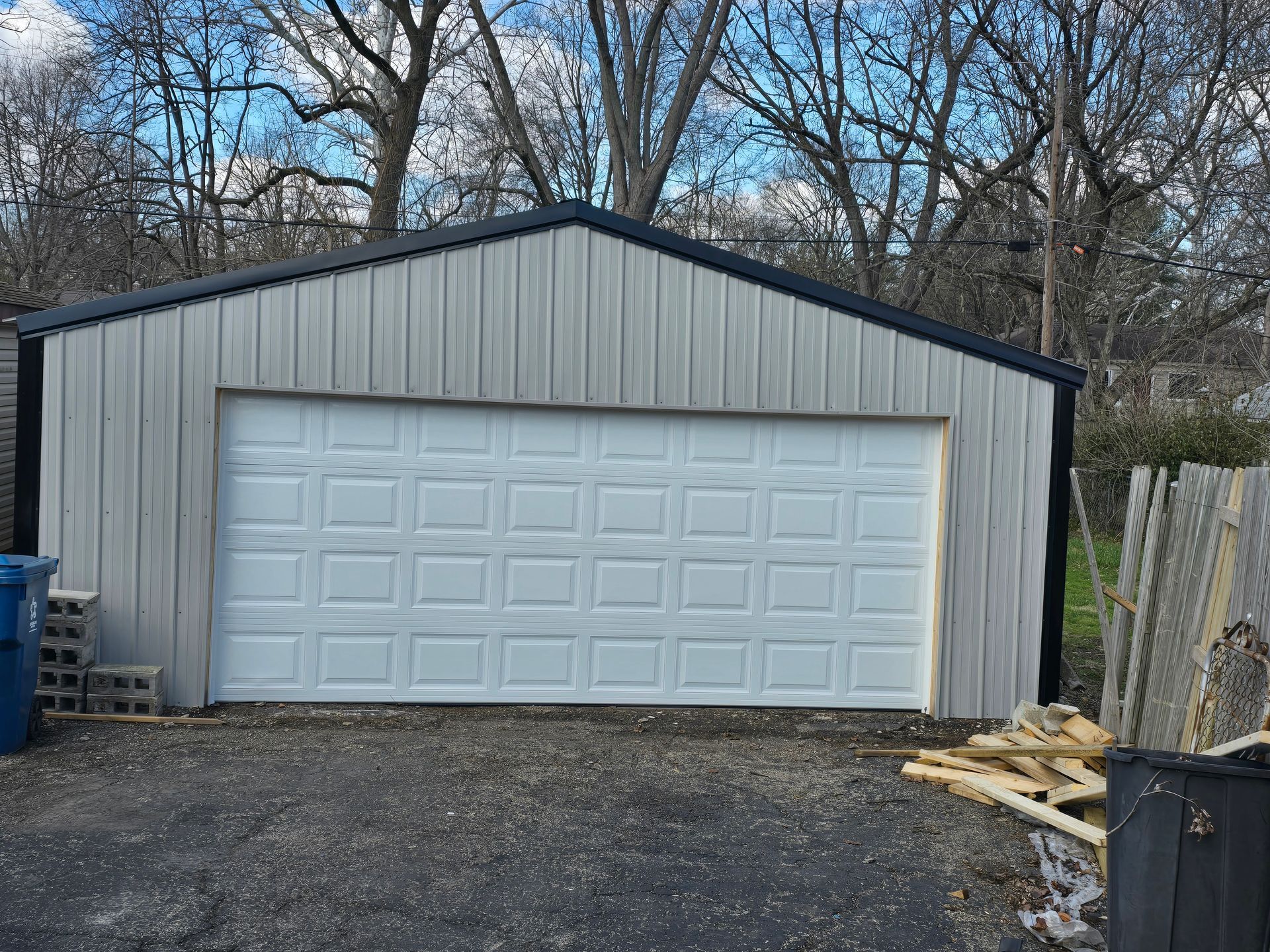 A detached garage with light grey metal siding and a white sectional garage door, set against a backdrop of bare trees.