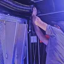 A person in a light shirt reaching up to adjust a dark, flexible vent hose inside an industrial enclosure.