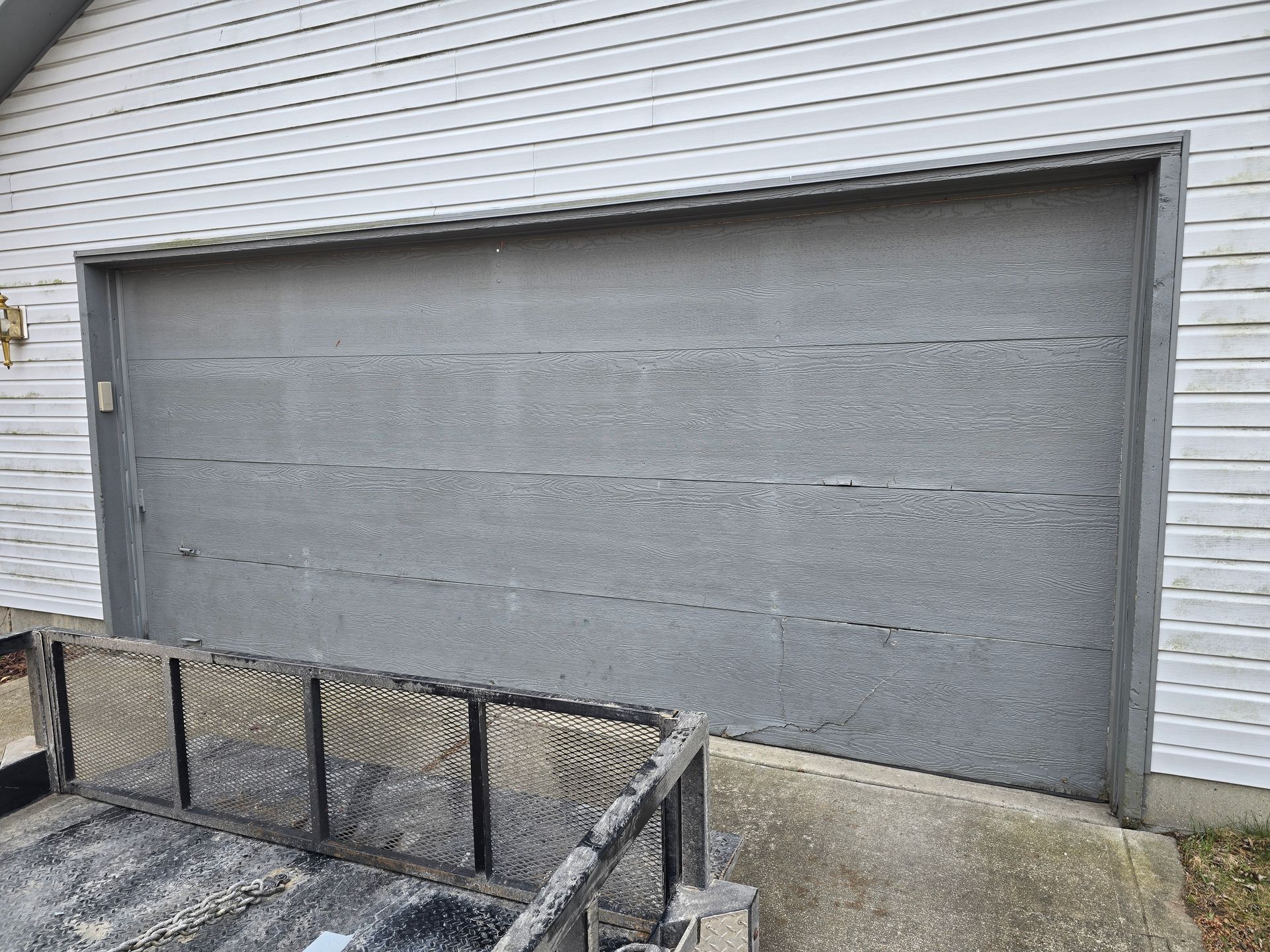 A gray garage door with horizontal panels set into a house with white horizontal siding and a trailer in the foreground.